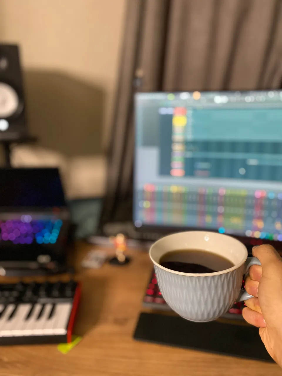 Elde bir fincan kahve; arkada bulanık stüdyo ekranı, MIDI klavye ve monitör